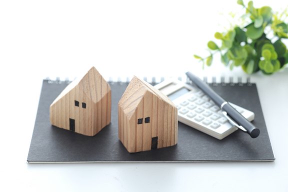 Zwei Modell-Holzhäuser stehen auf einer Mappe, auf der auch ein Taschenrechner und ein Stift liegen